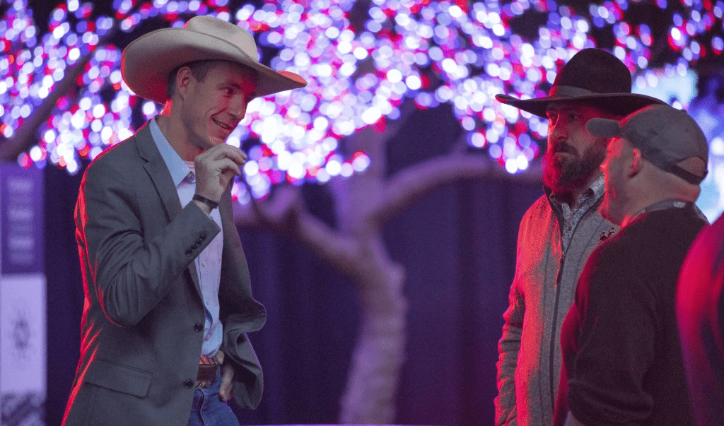 Three men wearing cowboy hats stand together talking under glowing purple and pink tree lights at a blockchain event. One man gestures with his hand while the others listen, creating a casual and festive atmosphere.