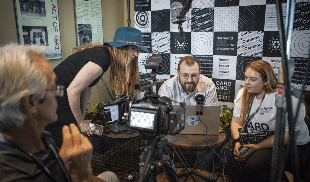 Four people are gathered around a laptop and camera setup, recording or livestreaming at a blockchain event. A patterned backdrop with words like “Cardano” and “impact” is behind them, along with visible cables and recording equipment.