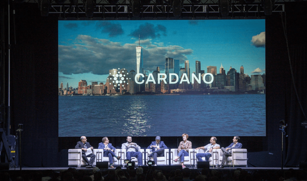 A panel of six people sit on stage at a blockchain event, in front of a large screen displaying the Cardano logo and a city skyline with water in the foreground.