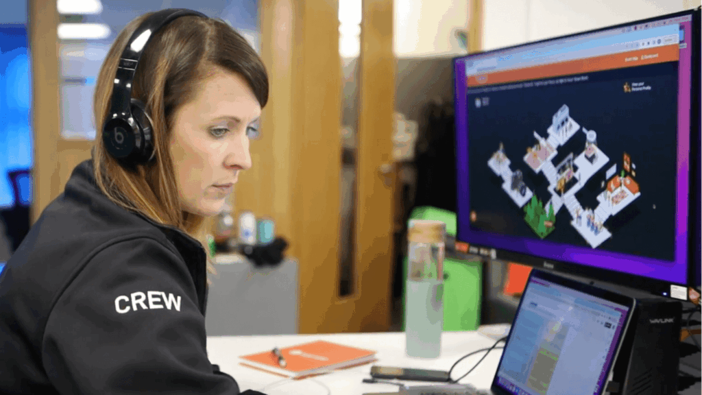 A woman wearing headphones and a jacket labeled CREW works at a desk with two monitors, one displaying a colorful diagram or map and the other showing text, during Just Eat Tech Fest 2021. A water bottle and notebook sit on her desk.