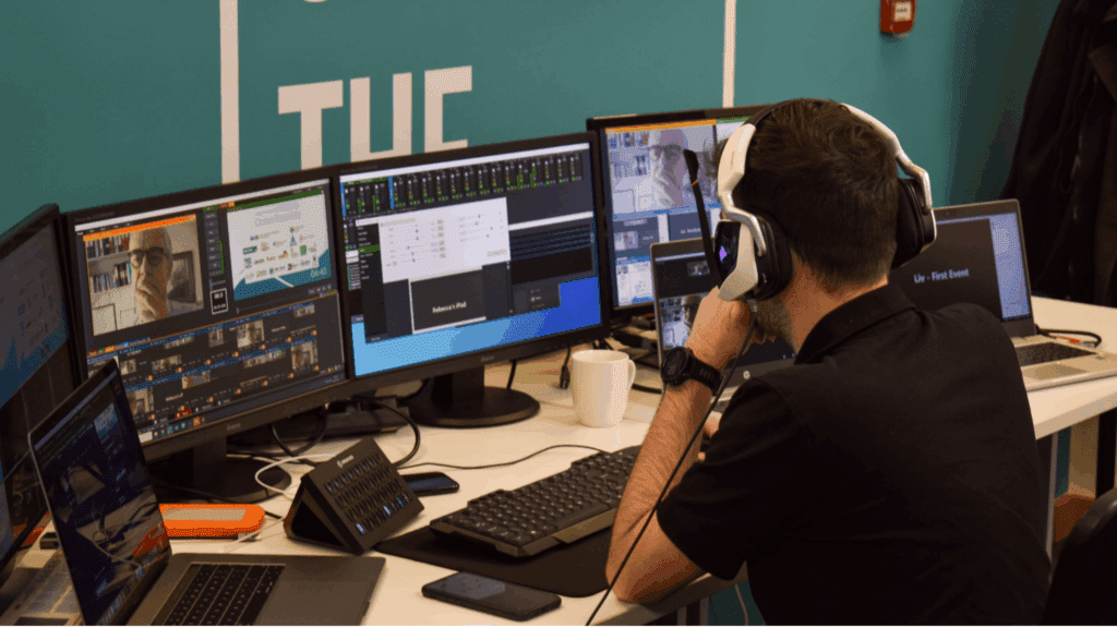 A person wearing headphones sits at a desk with multiple computer monitors displaying video calls and controls, suggesting they are managing a virtual global summit or coordinating a remote work or live streaming setup.