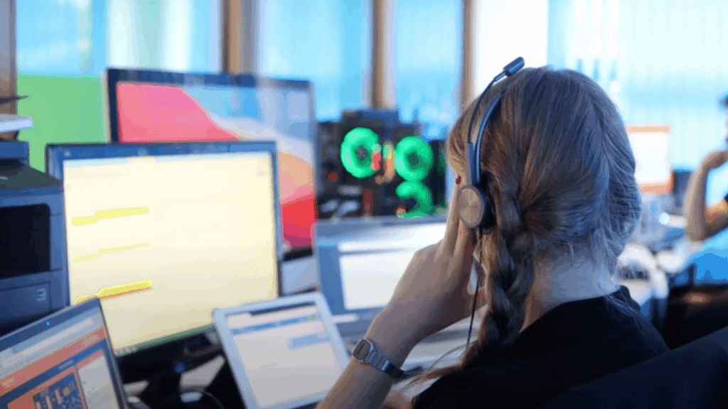 A person with long hair wearing a headset sits at a desk with multiple computer screens, appearing to coordinate a virtual global summit in a modern office or call center environment.