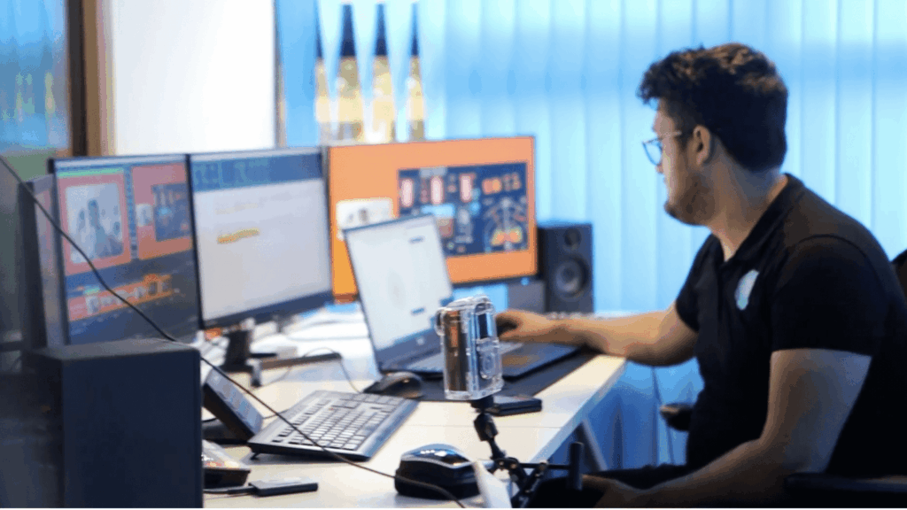 A man wearing glasses sits at a desk with multiple monitors, typing on a keyboard during a virtual global summit in a modern office with blue curtains and a microphone on the desk.
