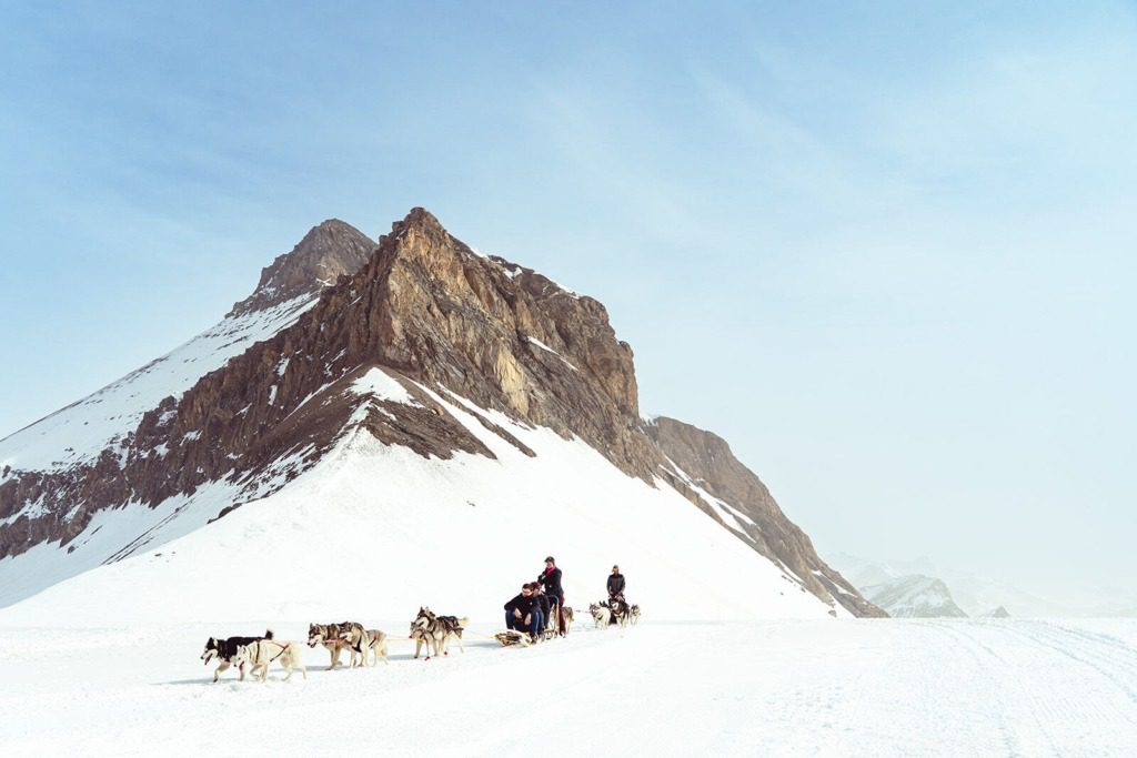Two people ride dog sleds pulled by huskies across a vast snowy landscape with a rugged, snow-covered mountain in the background under a clear sky.