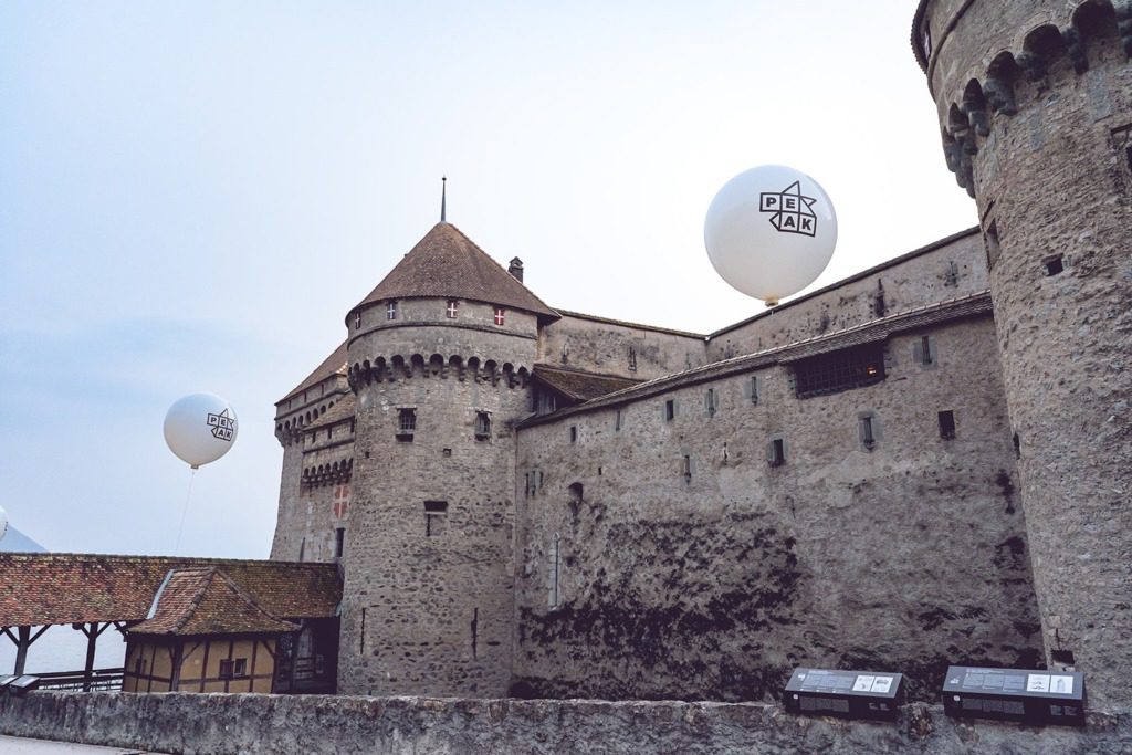 A stone castle with round towers and red-tiled rooftops stands beneath two large white balloons marked PEX TAK, floating under a clear sky. Informational signs in the foreground hint at an exclusive incentive travel event taking place.