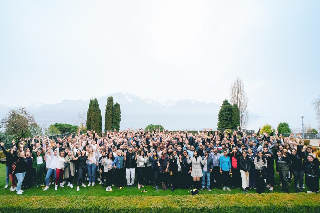 A large group of people pose together outdoors on a lawn, smiling and raising their arms, with trees and distant mountains visible in the background under a bright sky.