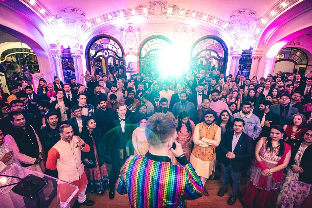 A large, diverse crowd stands in a decorated hall at an incentive travel event as a person in a colorful, rainbow-striped jacket addresses them from the front, with bright lights illuminating the ornate room.