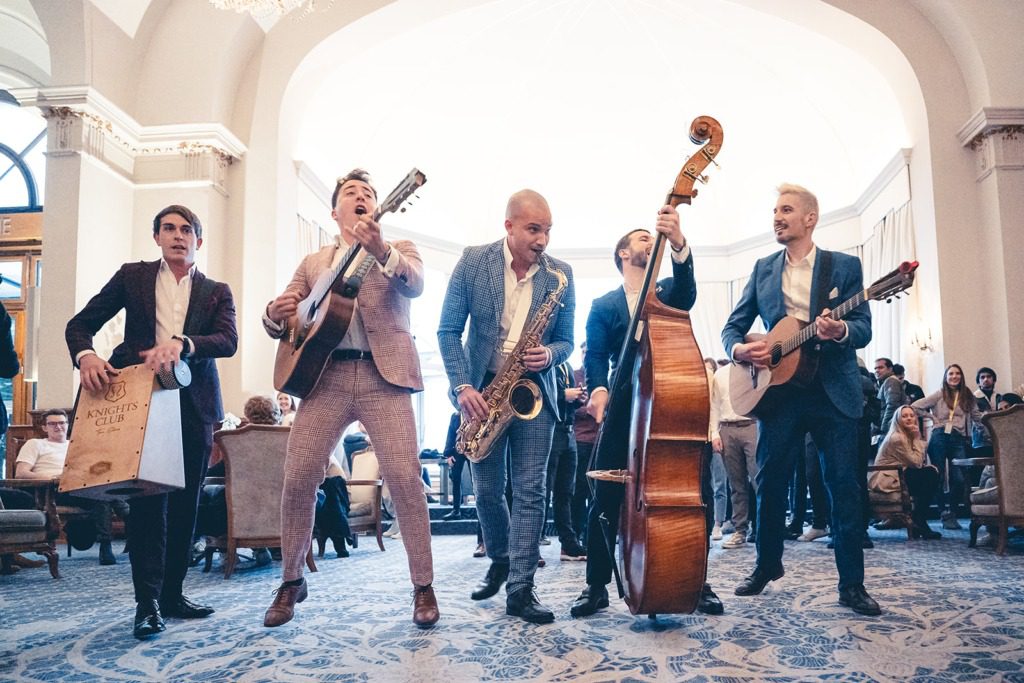 Five musicians in suits enthusiastically perform indoors at an incentive travel event, playing guitar, saxophone, upright bass, small guitar, and a box drum as the audience watches from seated chairs in a bright, elegant room.
