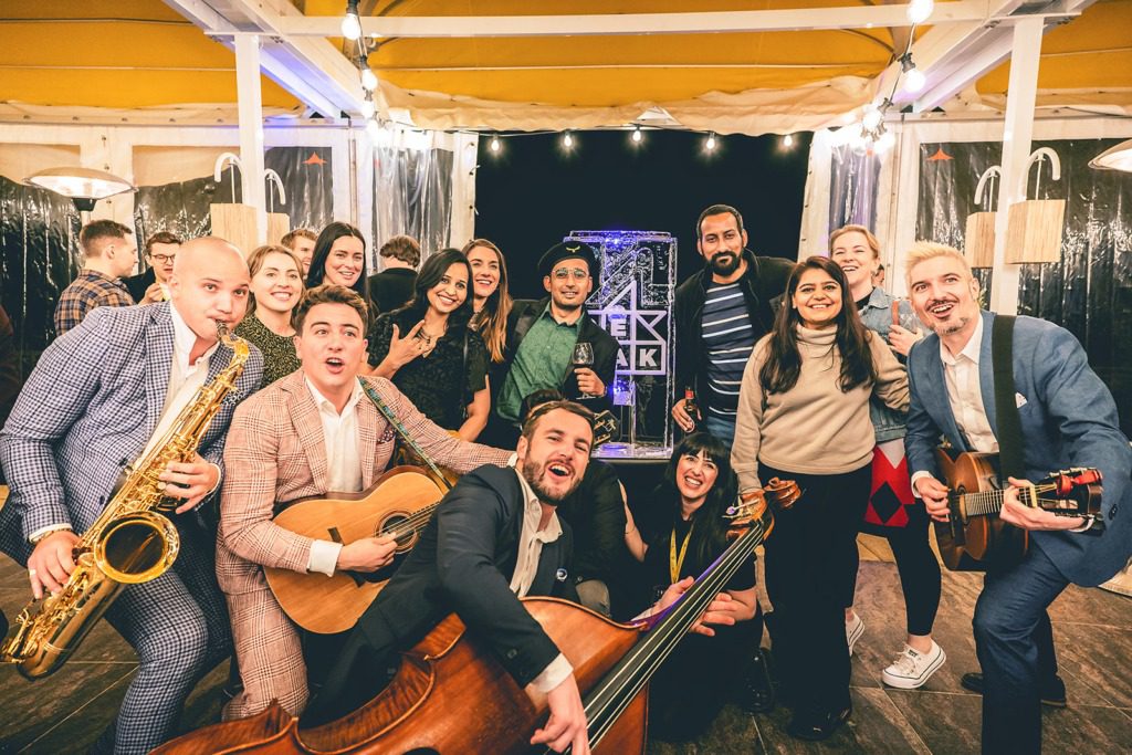 A group of people smiling and posing together indoors at an incentive travel event. Some are holding musical instruments including a saxophone, guitars, and a double bass. String lights and event decorations are visible in the background.