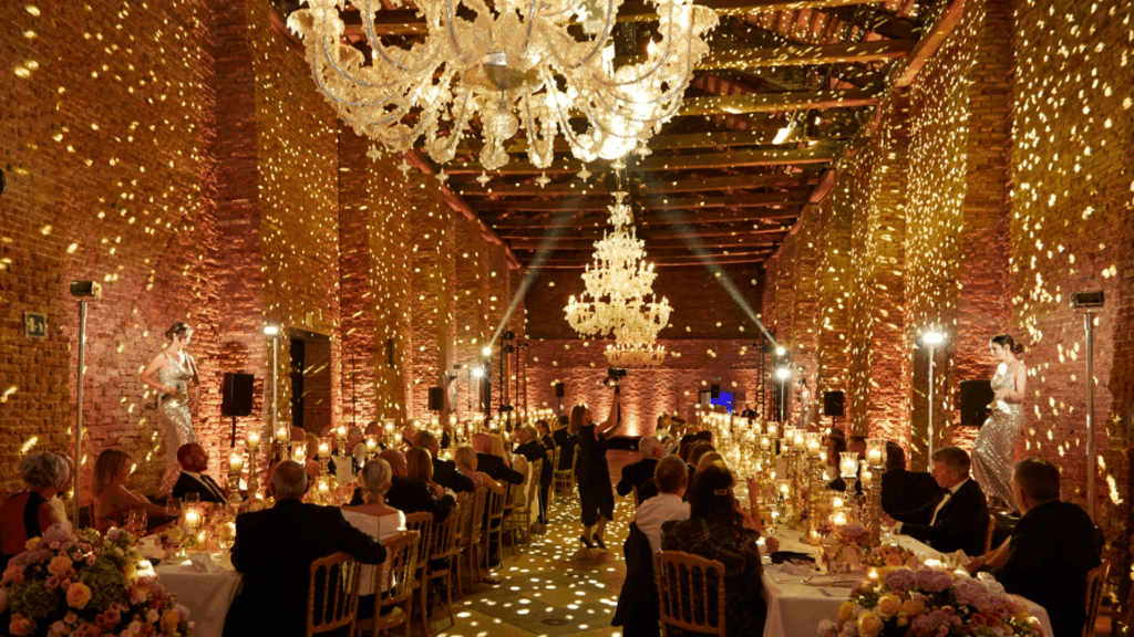A luxurious ballroom with exposed brick walls, chandeliers, and golden lights sets the scene for the Orient Express to Venice event, as guests at decorated tables watch a performer dance in the center, creating a festive, elegant atmosphere.
