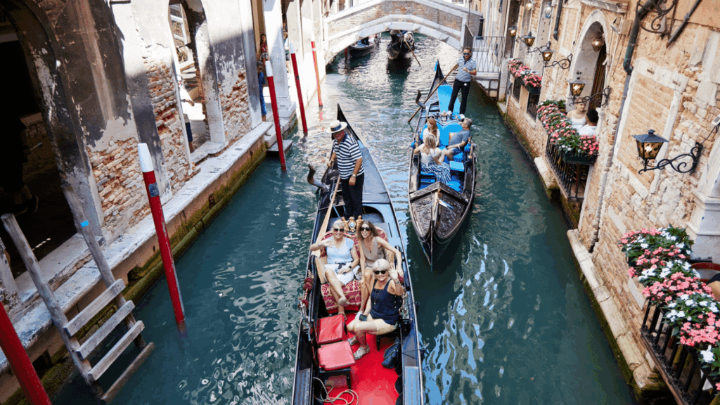 Gondolas with tourists glide through a narrow canal in Venice, Italy, as the city buzzes with excitement for the Orient Express to Venice event. A gondolier in a striped shirt rows beneath a small arched bridge amid historic brick buildings.