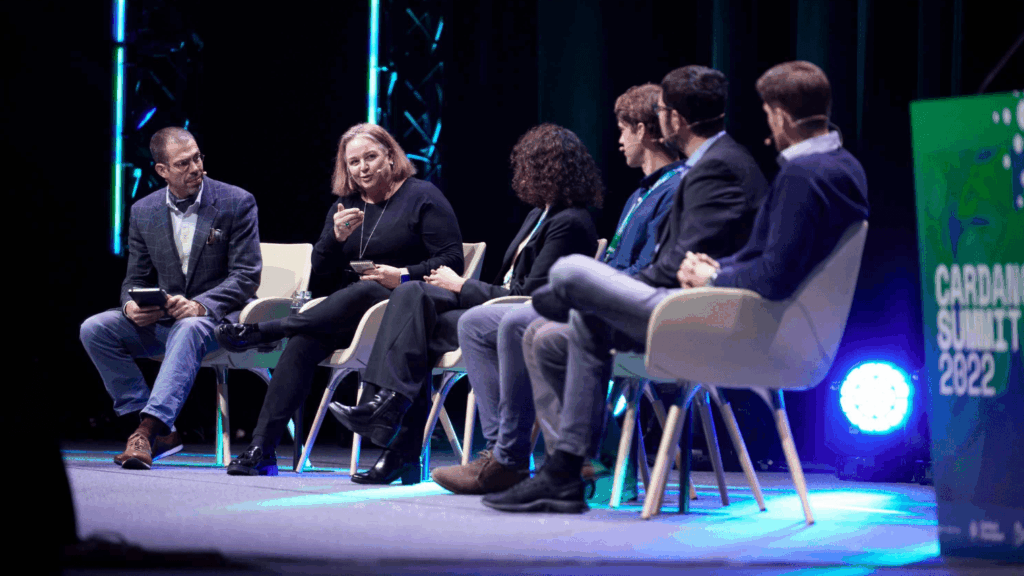 Six people are seated on stage in a panel discussion at the Cardano Summit. One woman is speaking while others listen. A sign in the foreground reads Cardano Summit 2022, with blue and green lighting highlighting the scene.