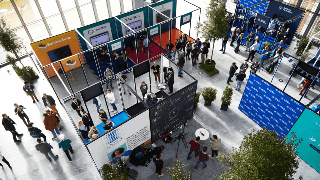 Aerial view of a busy convention hall during Cardano Summit, with multiple booths, groups of people networking, and large windows letting in natural light. Booths display company names like SoundSwap, Coinloan, and Taurus.