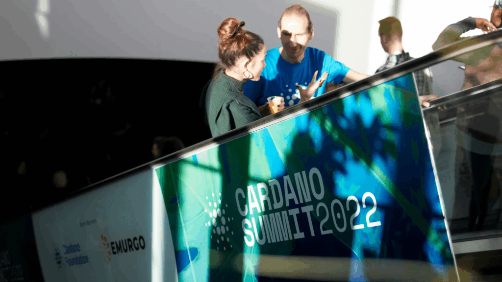 Two people talk beside a glass railing at this community event, with a sign reading Cardano Summit 2022. Sunlight casts shadows, while Cardano Foundation and EMURGO logos are visible on the panel.