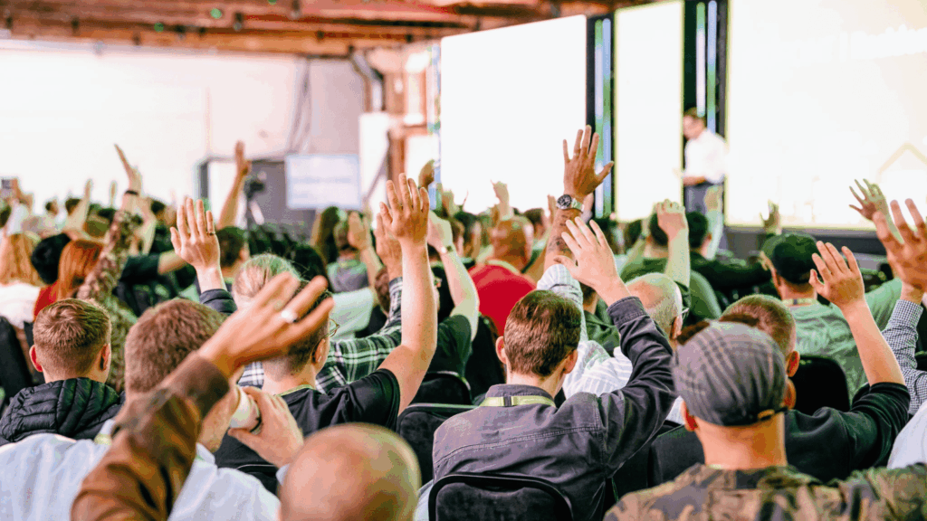A large group of people sit facing a stage, raising their hands in response to a speaker standing in front of bright screens during a sustainability & diversity conference or seminar.