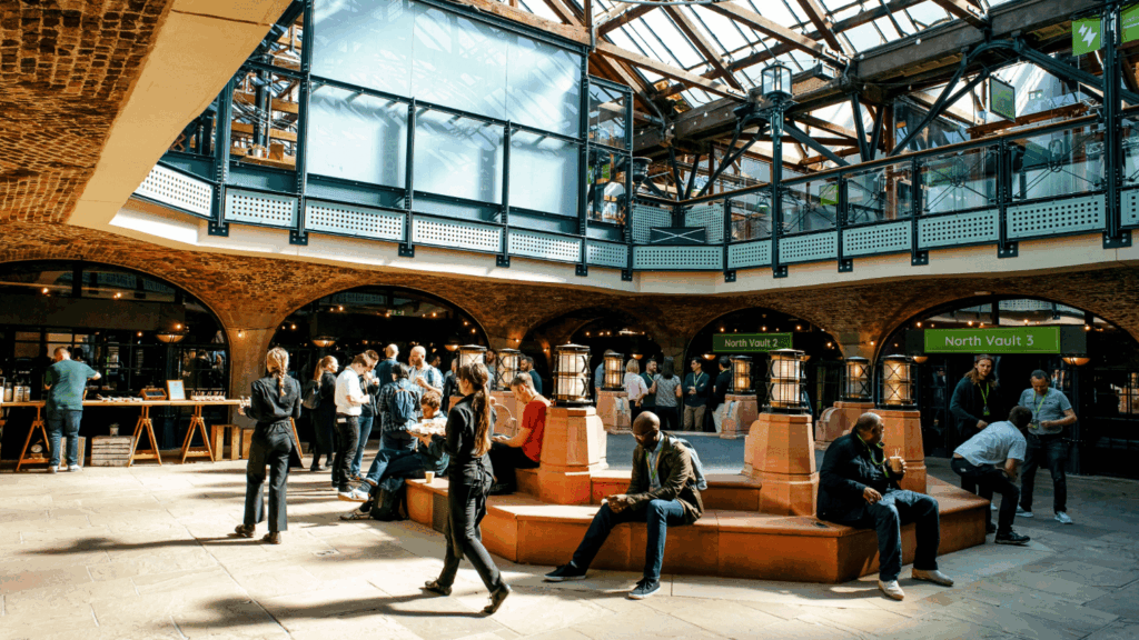 People stand and sit in a bright, spacious indoor atrium during a sustainability & diversity conference, surrounded by a glass ceiling, brick arches, and modern metal railings. Some converse in groups, while others use their phones or sit quietly on the central bench.