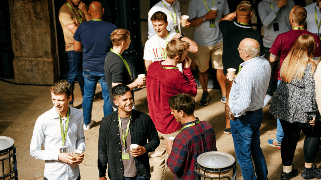 A group of people wearing name badges and holding cups gather indoors, chatting and smiling in small groups at a sustainability & diversity conference, suggesting a casual networking or social event.