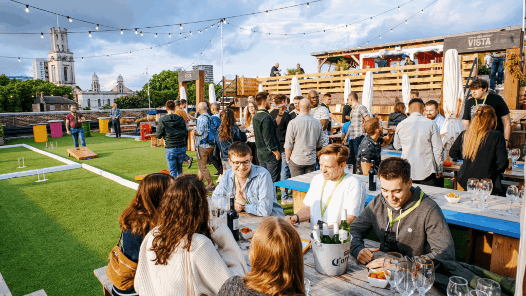 A group of people enjoy drinks and conversation at outdoor wooden tables on a rooftop bar, networking after the sustainability & diversity conference, with string lights overhead and city buildings and a church tower in the background.