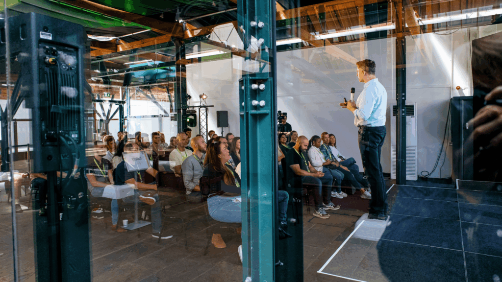 A man stands on a stage speaking into a microphone to an audience at a sustainability & diversity conference, inside a modern industrial-style building with exposed beams and glass walls.