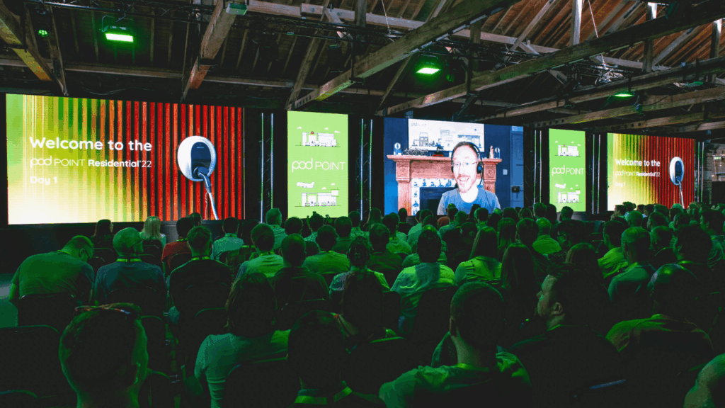 A large audience sits in a dimly lit hall watching multiple screens displaying a welcome message for the “podPOINT Residential ’22” sustainability & diversity conference, while a speaker joins via video call amid vibrant green lighting.