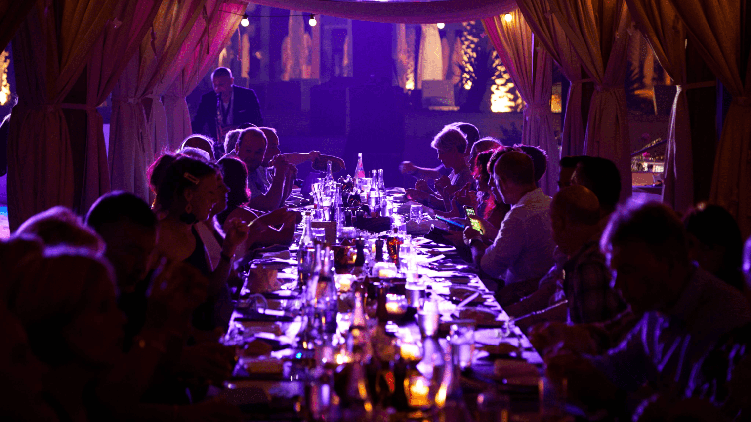 A large group of people sits around a long, candlelit table under a canopy at night, sharing a meal and drinks in a warm, festive atmosphere with purple lighting—an unforgettable Platinum Club gathering.