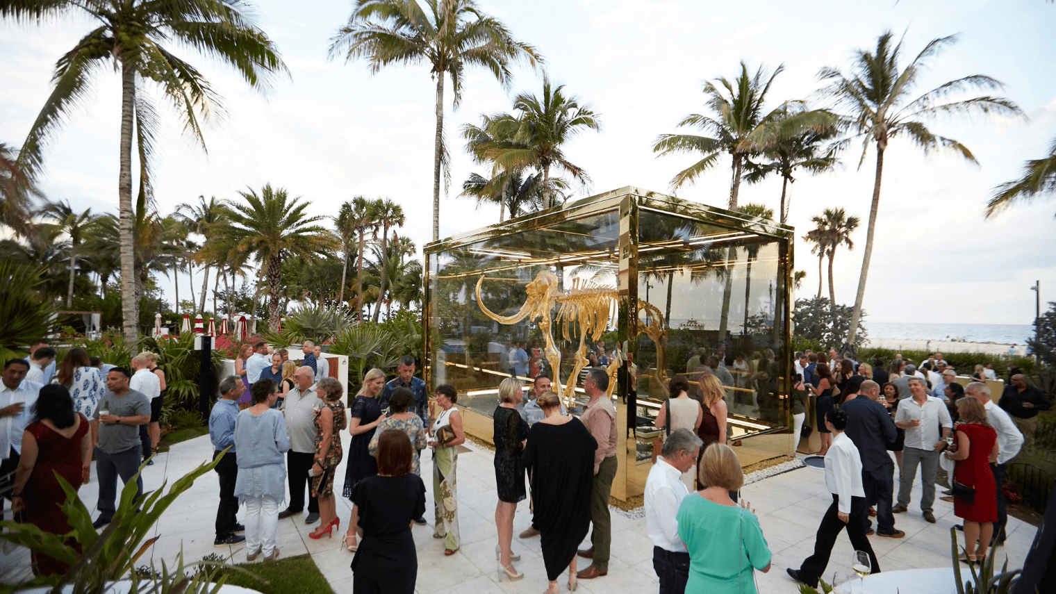 A large group gathers outdoors around a golden glass enclosure containing a golden animal skeleton at the Platinum Club, surrounded by palm trees and overlooking the ocean. The event appears formal and festive.