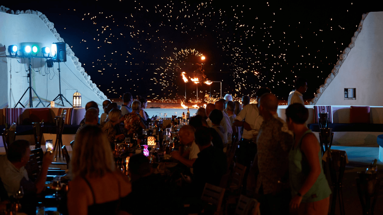 A crowd of people seated at outdoor tables watch a fire performer at night, sparks and flames lighting up the dark sky. The festive event, hosted by the Platinum Club, glows with colorful lights and decorations.