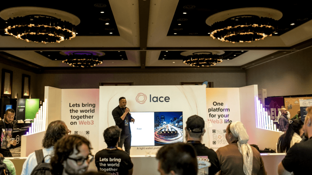 A modern exhibition stand for Lace features a central screen showing a man’s face, white walls with branded text, neon light decorations, and neatly stacked black t-shirts on both sides. The setting appears bright, welcoming, and expresses a subtle thank you to visitors.