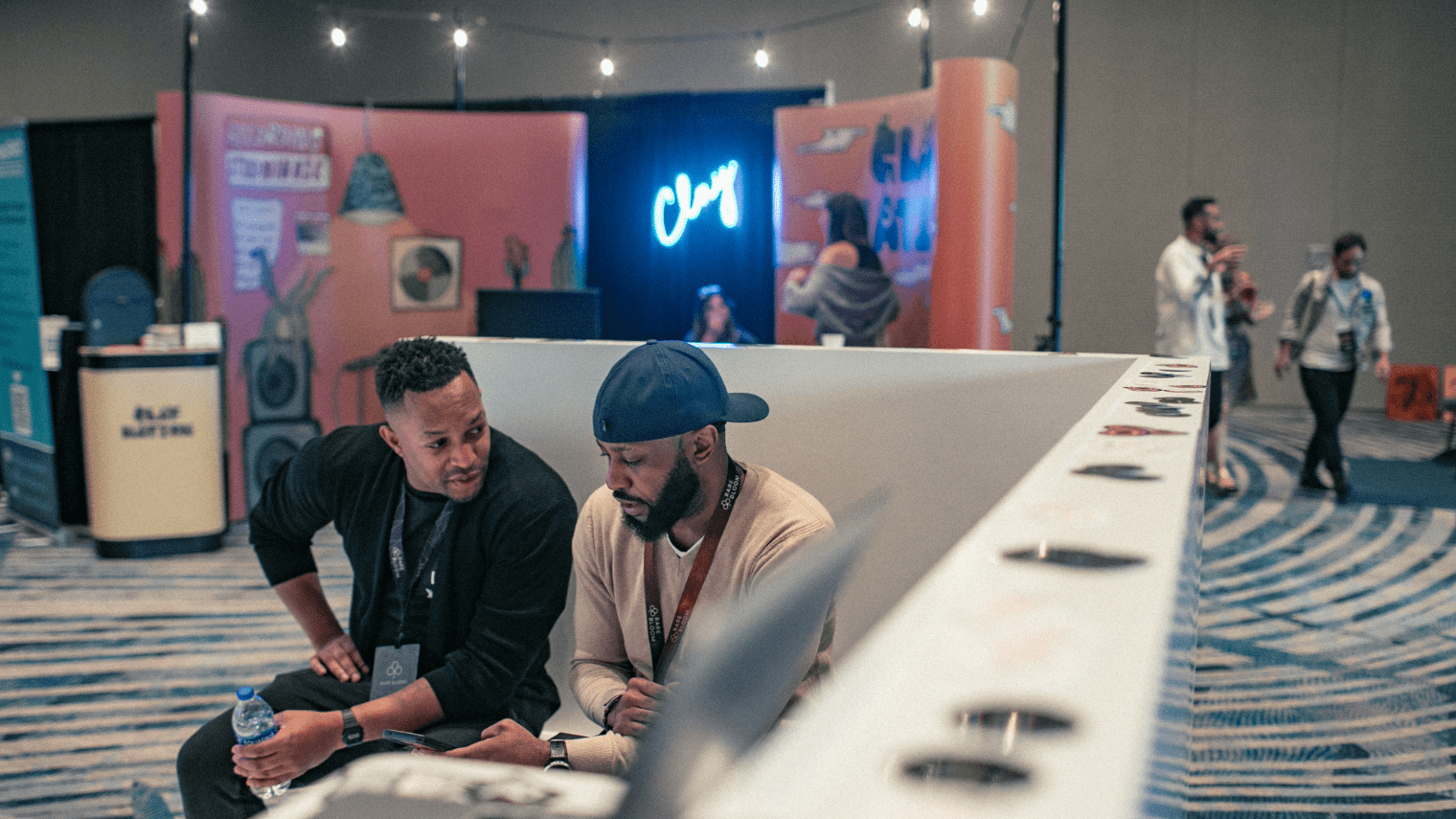 Two men sit and talk at an experiential marketing event booth with colorful displays and a neon “Chuy” sign in the background, while others mingle in the lively event space.