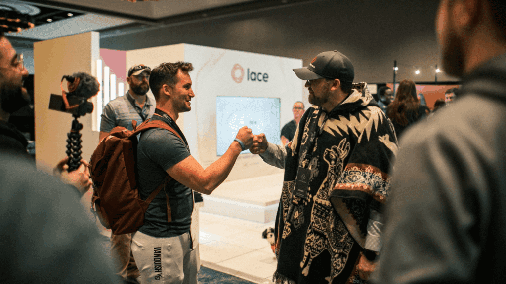 Two men greet each other with a fist bump at an experiential marketing event, surrounded by people and camera equipment. One sports a backpack and casual clothes, while the other wears a patterned poncho and cap. A booth labeled lace is in the background.