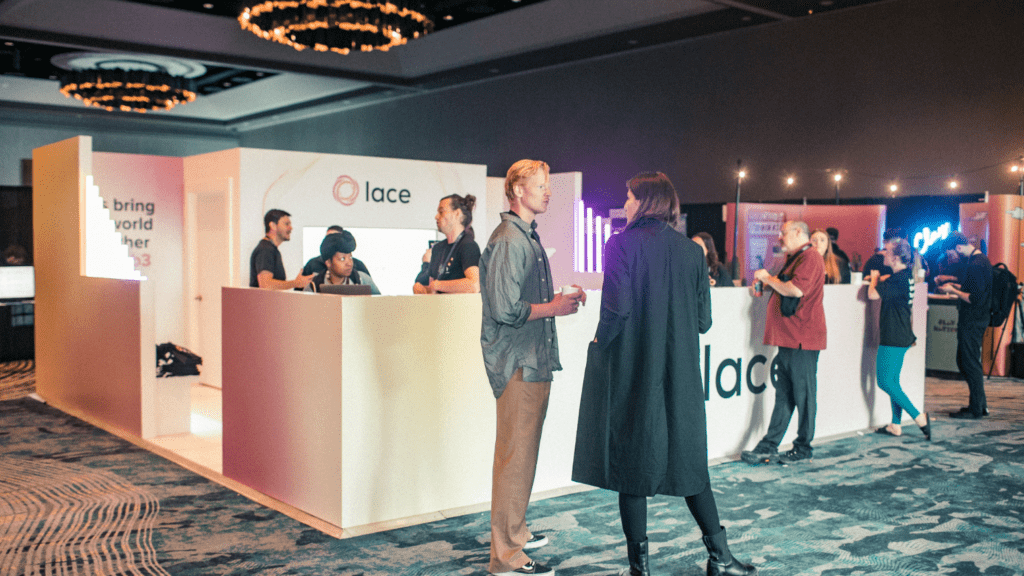 A modern exhibition stand for Lace features a central screen showing a man’s face, white walls with branded text, neon light decorations, and neatly stacked black t-shirts on both sides. The setting appears bright, welcoming, and expresses a subtle thank you to visitors.