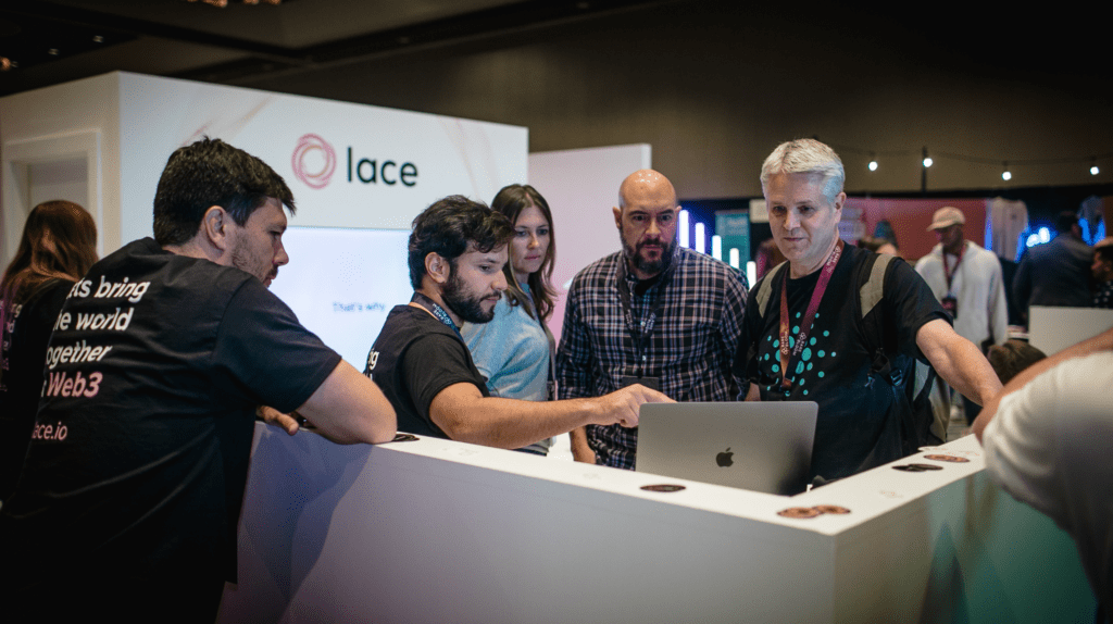 A group of five people gathers around a booth at a tech event, engaging in experiential marketing as one man points at a laptop screen while others watch attentively. A “lace” sign and various tech displays are visible in the background.