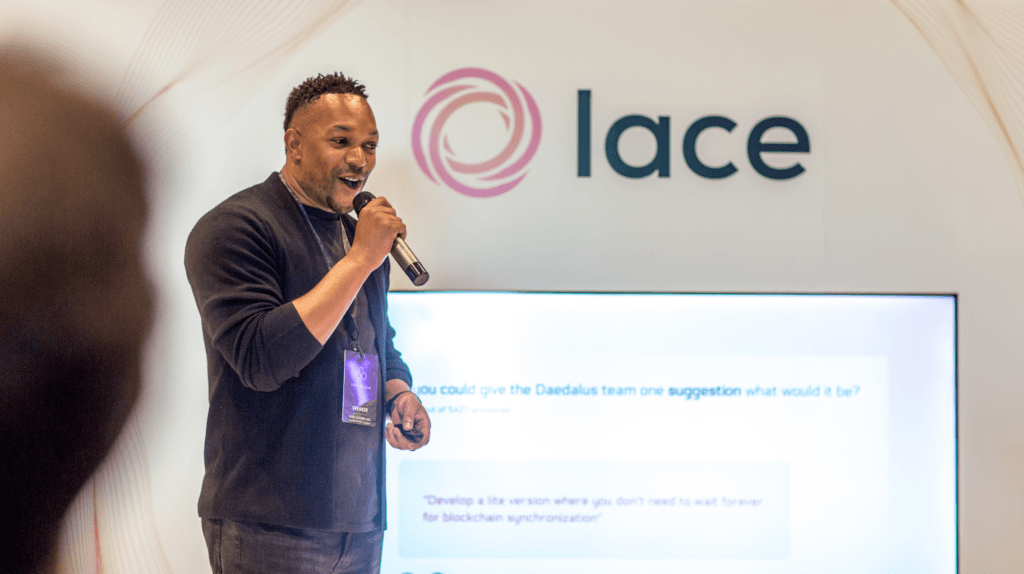 A man speaks into a microphone on stage in front of a screen and a sign that reads lace. Wearing a dark sweater and lanyard, he appears to be presenting on experiential marketing at an event or conference.