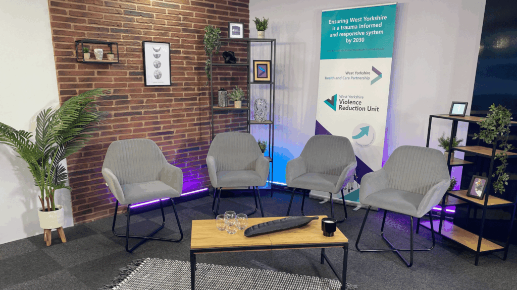 Four gray chairs are arranged in a semi-circle around a wooden coffee table with glasses and a tray, set in a cozy studio with decorative plants, shelves, and a Knowledge Exchange banner about West Yorkshire’s trauma-informed response system.