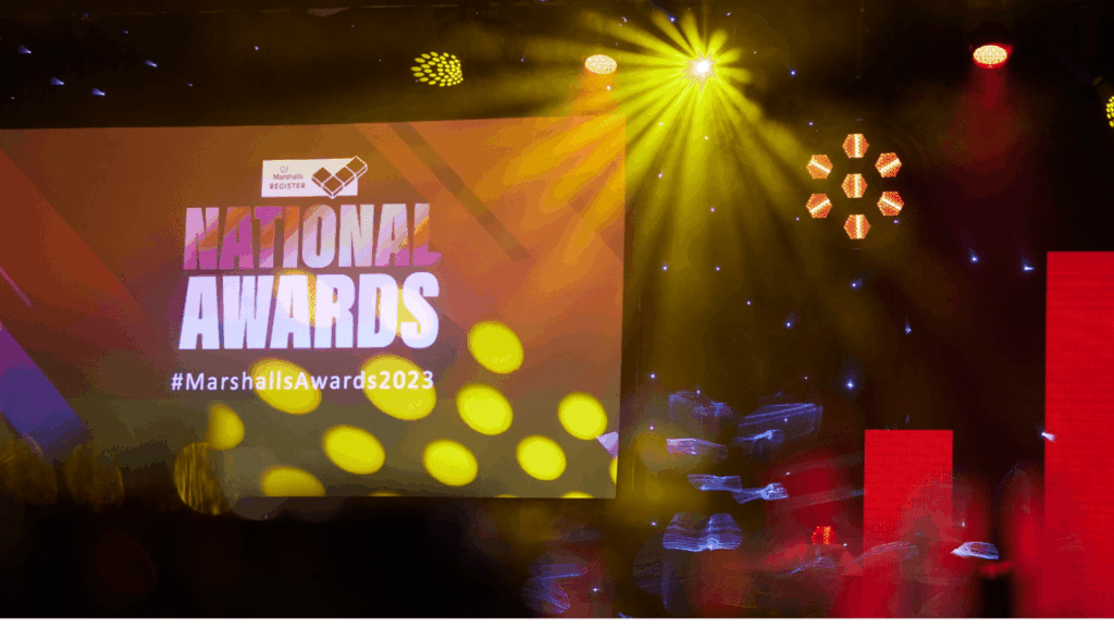 A stage with colorful lights and a large screen displaying NATIONAL AWARDS and #MarshallsAwards2023 during an electrifying awards roadshow event.