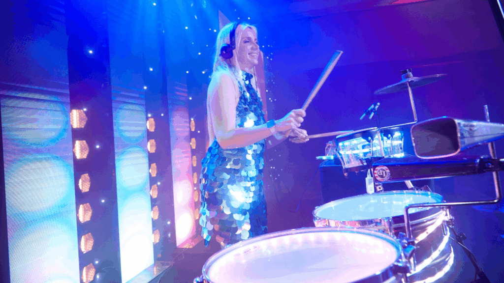 A woman in a sequined dress plays electronic drums onstage under blue and purple lights at an awards roadshow, smiling and wearing headphones.