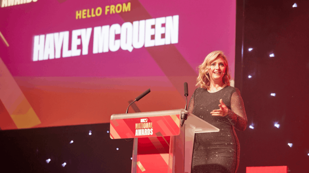 A woman stands at a podium on stage, speaking and gesturing with one hand. Behind her, a large screen displays HELLO FROM HAYLEY MCQUEEN at a Roadshow and Awards event.