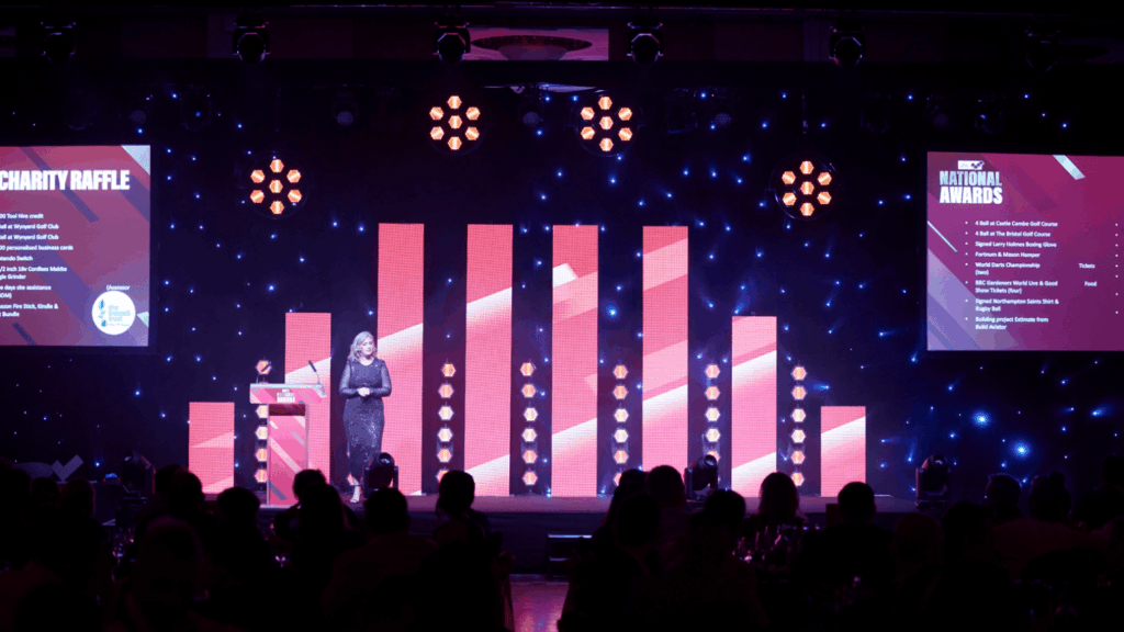 A woman stands on stage at a Roadshow and Awards event, framed by vertical red lights and spotlights. Two large screens display charity raffle and national awards details as audience members watch from the foreground.