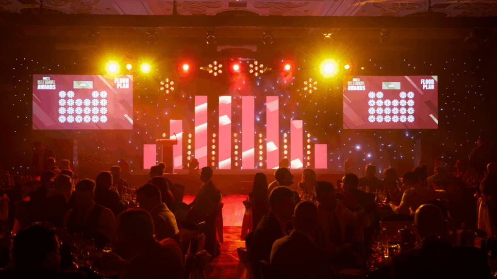 A dimly lit Roadshow and Awards ceremony room with a stage featuring tall, illuminated columns, orange and yellow spotlights, two large screens displaying text, and an audience seated at round tables.