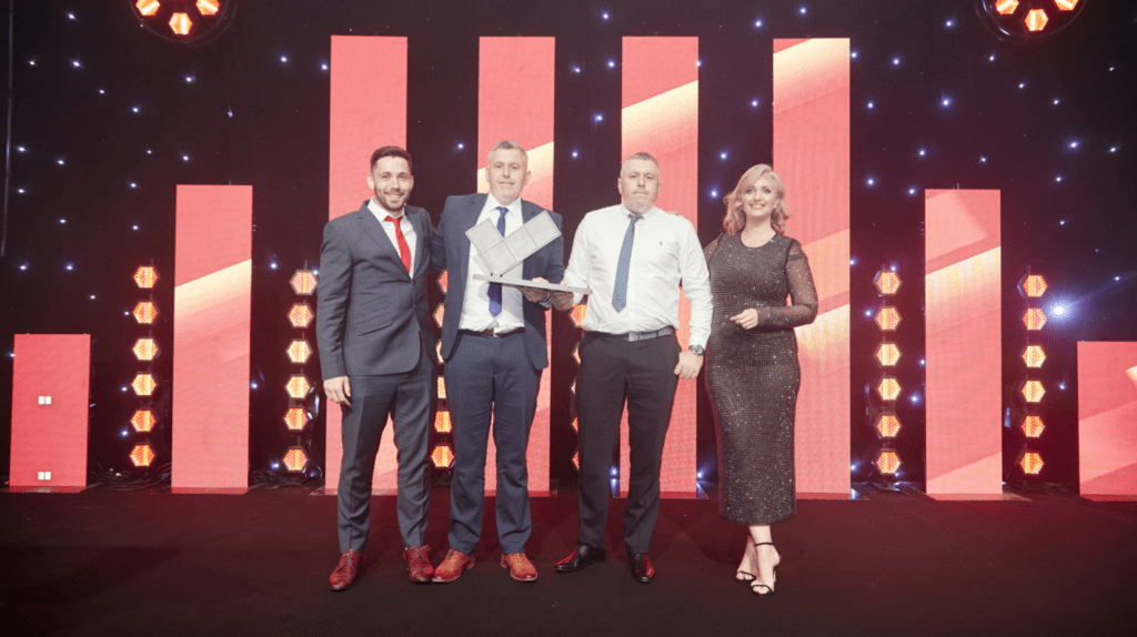 Four people stand on a stage in formal attire, holding an award at a prestigious awards roadshow. The background features vertical red-lit panels and star-like lights, creating a celebratory atmosphere.