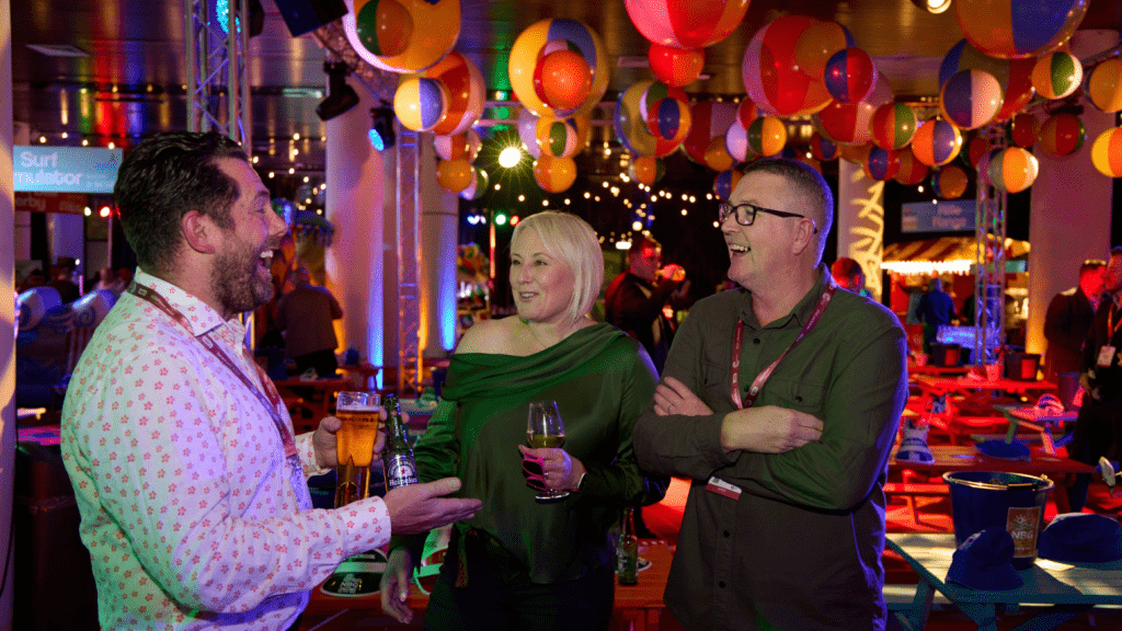 Three people stand together laughing and talking at a lively indoor conference & exhibition decorated with colorful balloons and string lights; each holds a drink, and tables with drinks and snacks are visible in the background.