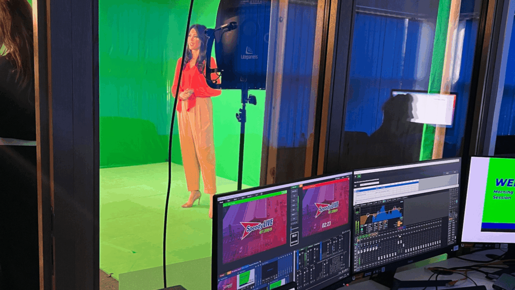 A woman stands in front of a green screen in a studio, preparing for a Net Zero Webinar as video production monitors display broadcast controls and graphics in the foreground.