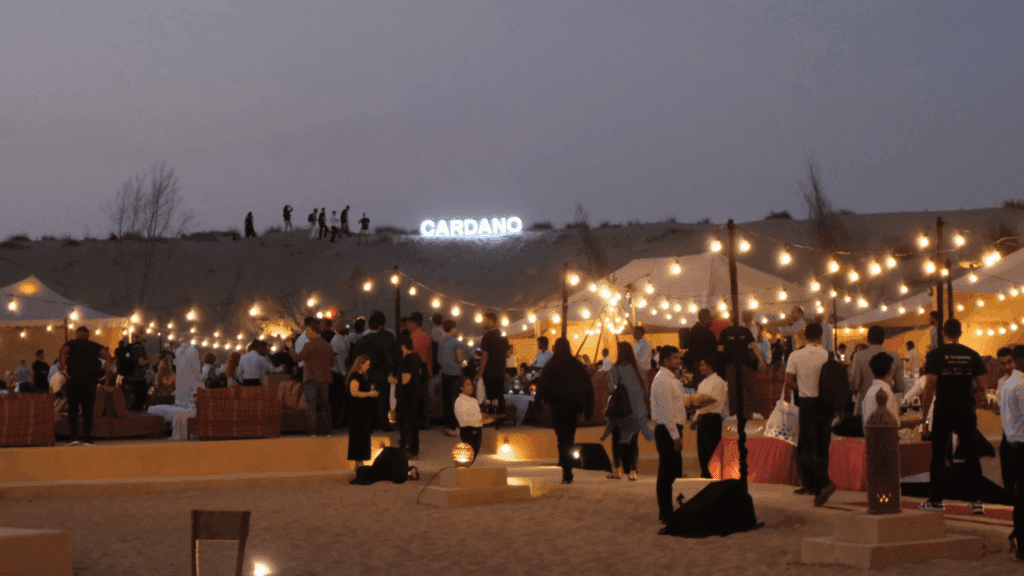 A lively outdoor evening event in the desert with string lights, people socializing, and a glowing CARDANO sign on a sand dune in the background. Tents and seating areas set the scene for Cardano Summit 2023 under the night sky.