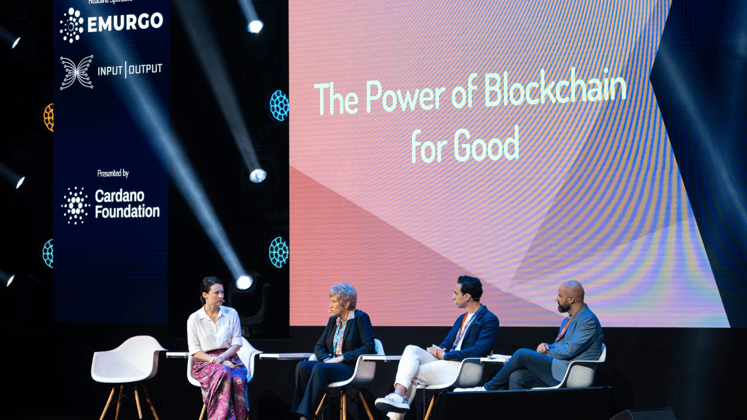 Four panelists sit on stage under bright lights at Cardano Summit 2023, discussing The Power of Blockchain for Good. Logos for EMURGO, Input Output, and Cardano Foundation are displayed on the backdrop.