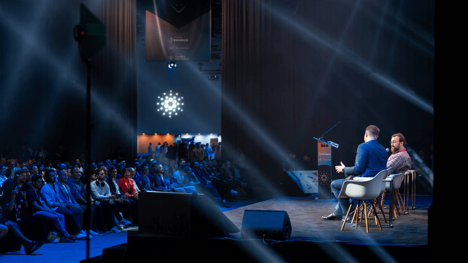 Two speakers sit on stage, facing an audience at a large indoor event during Cardano Summit 2023. Bright spotlights shine across the room, highlighting the speakers and creating a dynamic atmosphere as the audience watches attentively.