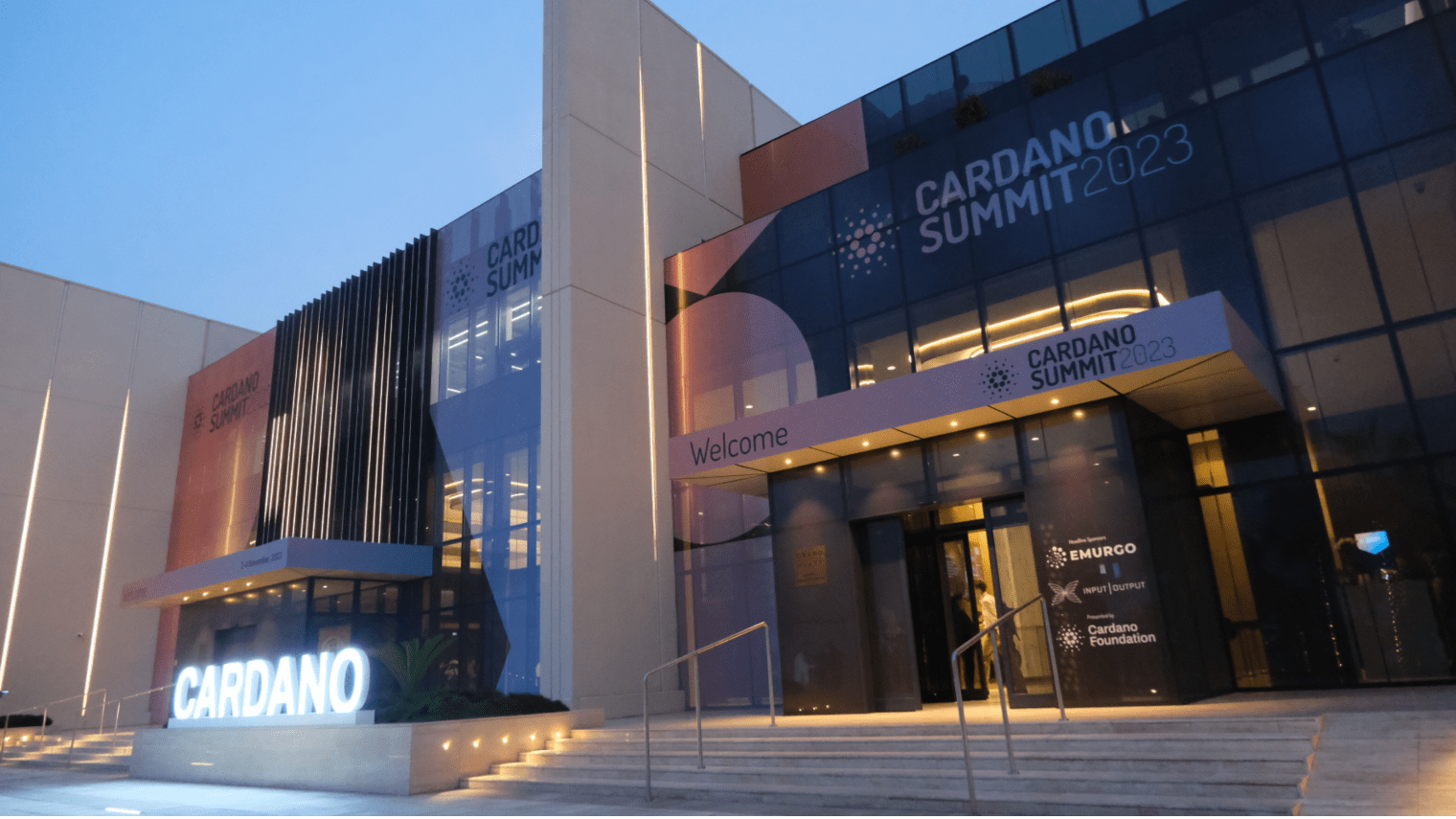 Modern building with Cardano signage and banners for Cardano Summit 2023. Lit steps lead to glass doors; the event welcomes participants to the Cardano Summit 2023. Exterior is illuminated in the early evening.