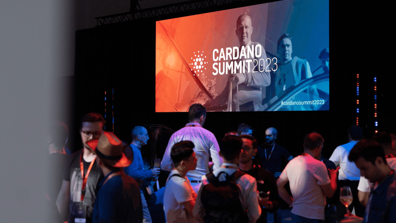 A group socializes at an indoor conference, gathered before a large screen showcasing “Cardano Summit 2023” visuals and two men’s images, highlighting the excitement of the Cardano Summit 2023 event.