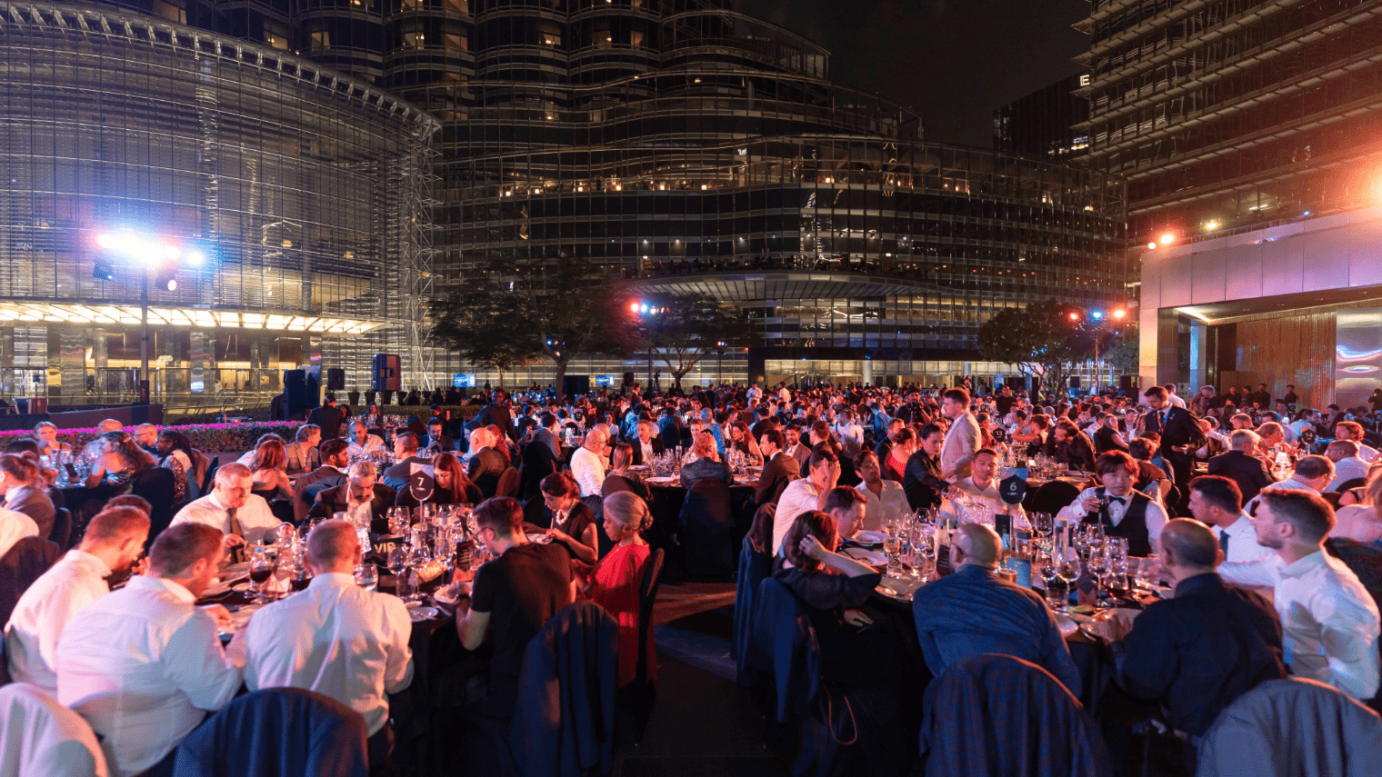 A large crowd dressed formally is seated at round tables outdoors at night, dining and socializing under bright lights during the Cardano Summit 2023, with modern glass buildings in the background.