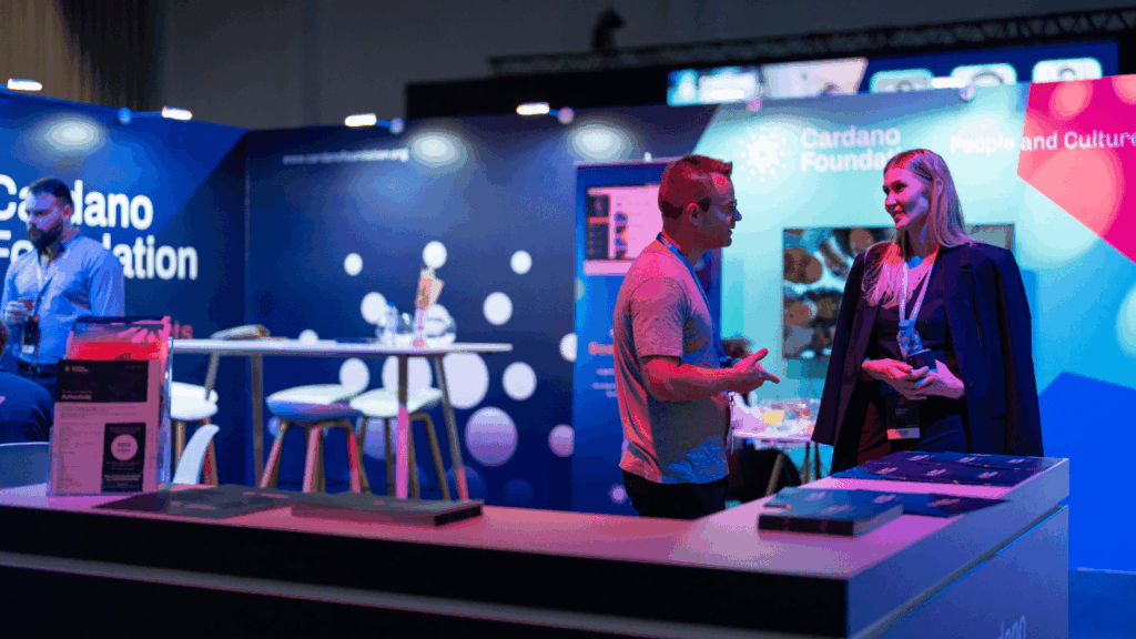 Two people talk at a booth with “Cardano Foundation” branding during Cardano Summit 2023. The area is lit with blue and purple lights, with brochures on the counter, stools, and a display in the background.