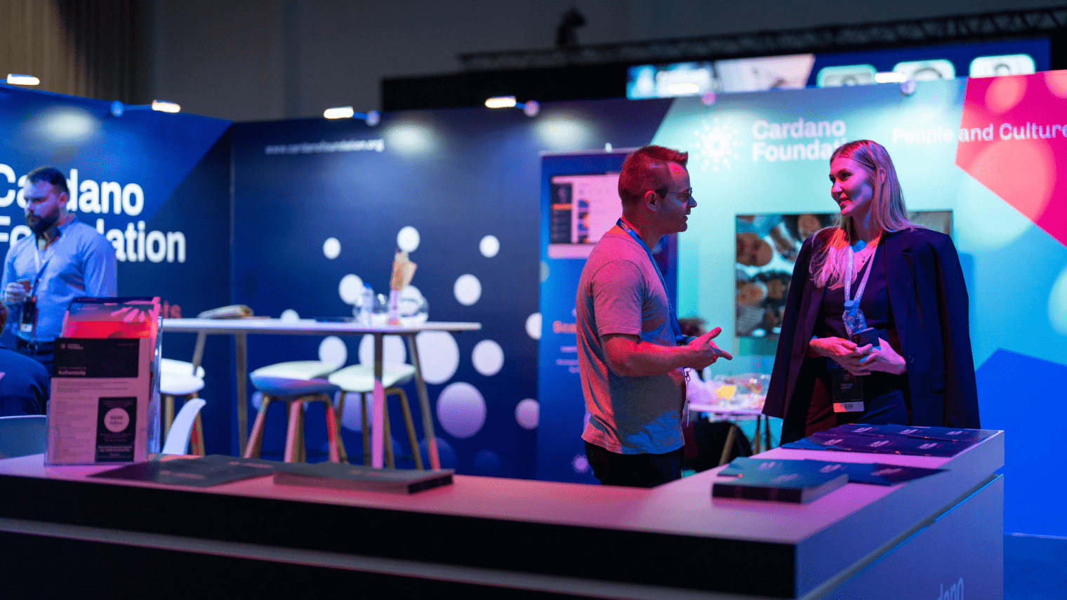 Two people talk at a booth with “Cardano Foundation” branding during Cardano Summit 2023. The area is lit with blue and purple lights, with brochures on the counter, stools, and a display in the background.